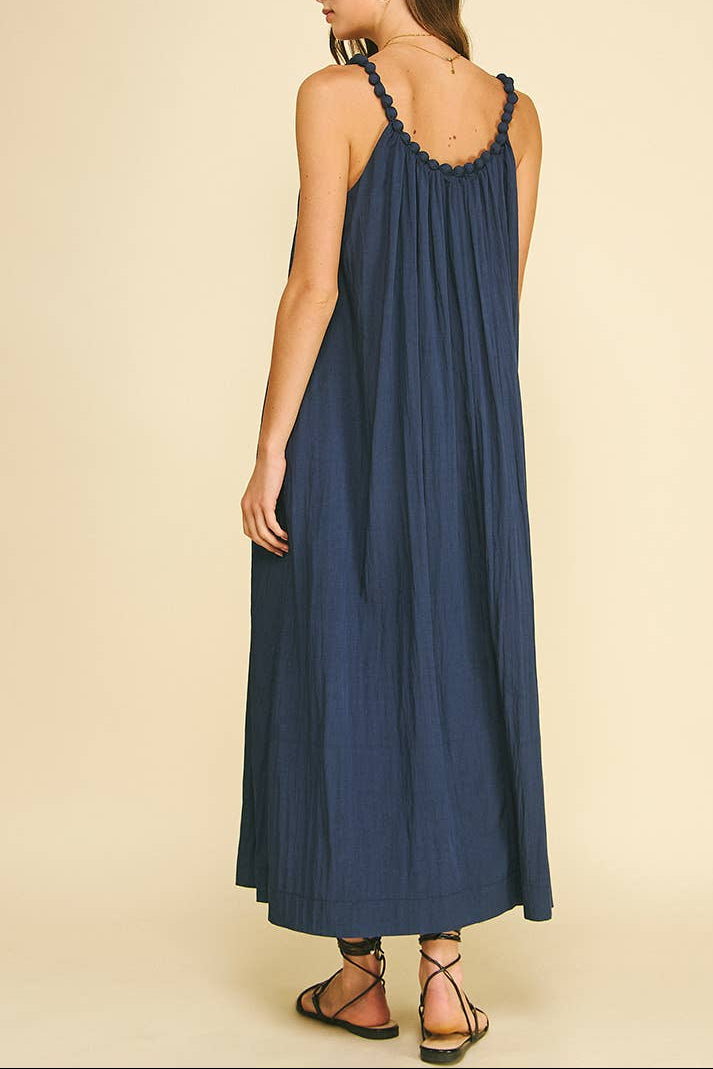 A woman modeling a sleeveless navy blue maxi dress from the back, featuring ruffled shoulder straps and a flowing pleated skirt, set against a solid beige background.
