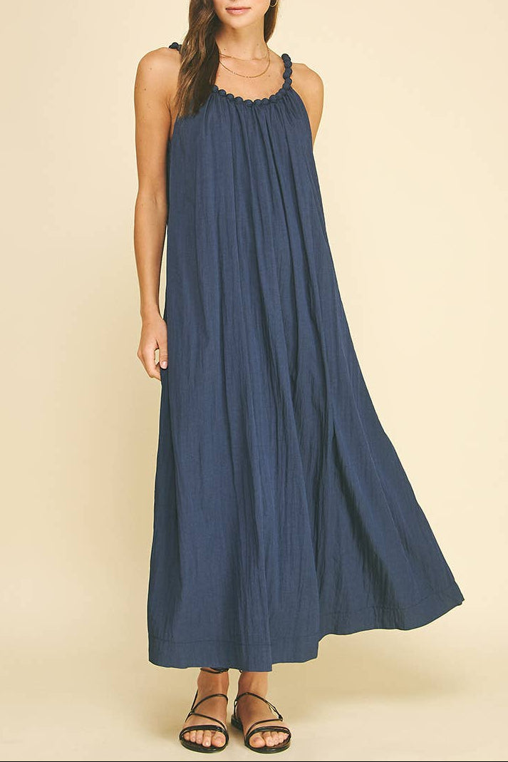 A woman modeling a sleeveless, floor-length navy blue maxi dress with a gathered neckline and flowing skirt, paired with brown strappy sandals against a solid beige studio background.