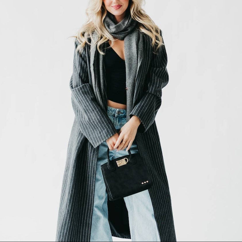 Woman wearing a long gray coat with a black scarf and holding a black handbag on a white background