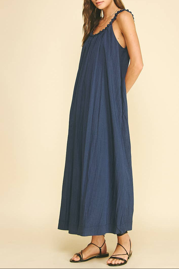 A woman modeling a sleeveless navy blue maxi dress with scalloped shoulder trim and black strappy flat sandals, set against a plain beige studio background.