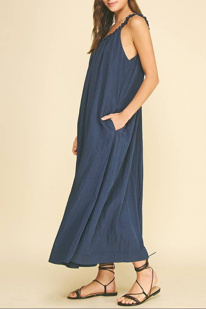 A woman modeling a sleeveless navy blue maxi dress with side pockets and a gathered neckline, paired with black strappy flat sandals, against a solid beige studio background.